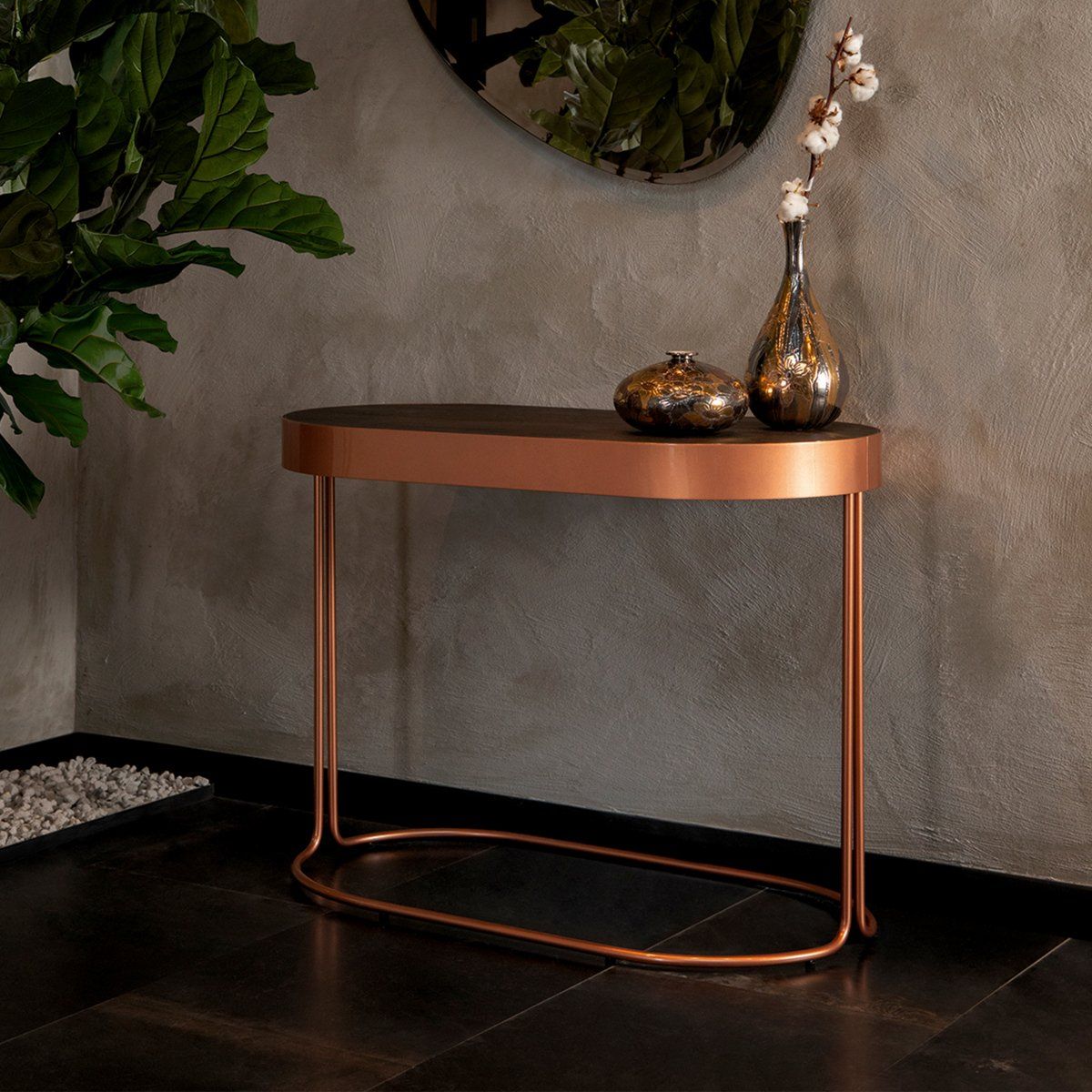 Una moderna consolle ovale con piano scuro e struttura in rame metallizzato, su cui sono appoggiati due vasi decorativi su una parete con finitura materica.