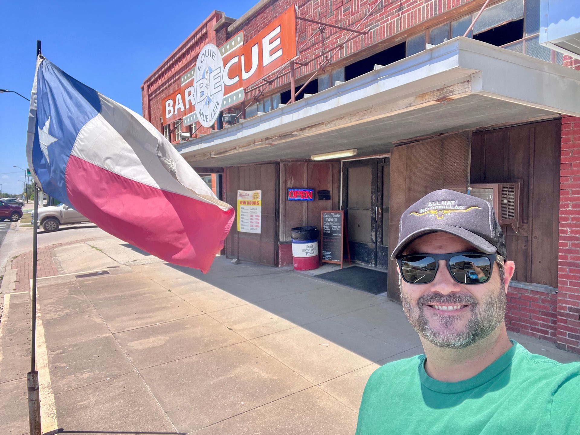 A man wearing sunglasses and a hat is standing in front of a barbecue restaurant.