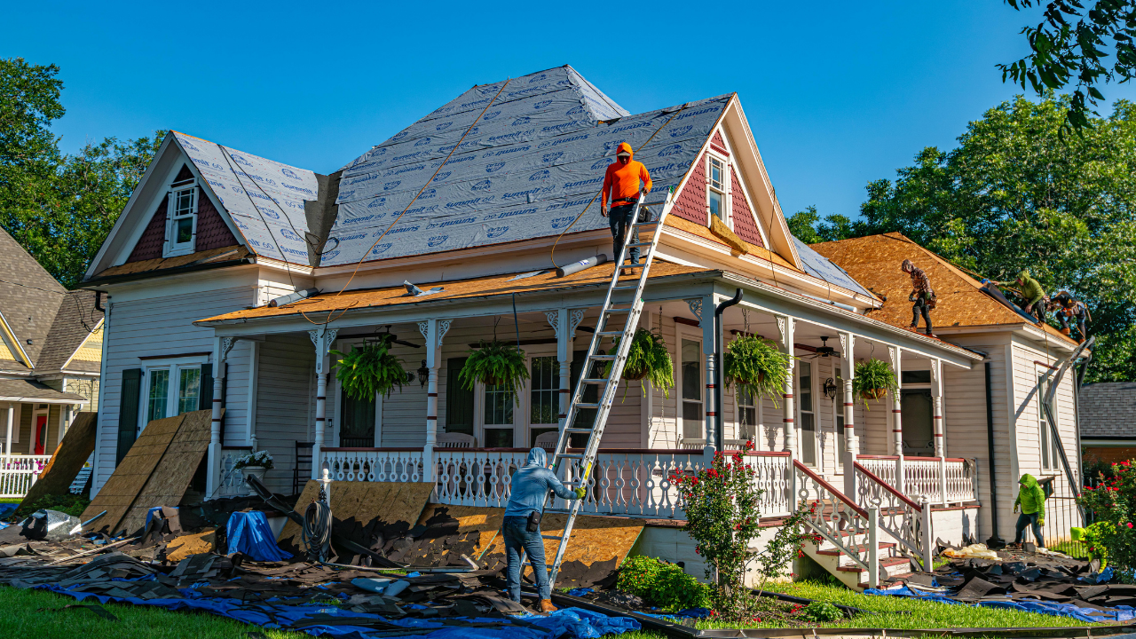 Workers replace a roof on a white house, with one worker on a ladder and another working on the roof underlayment.