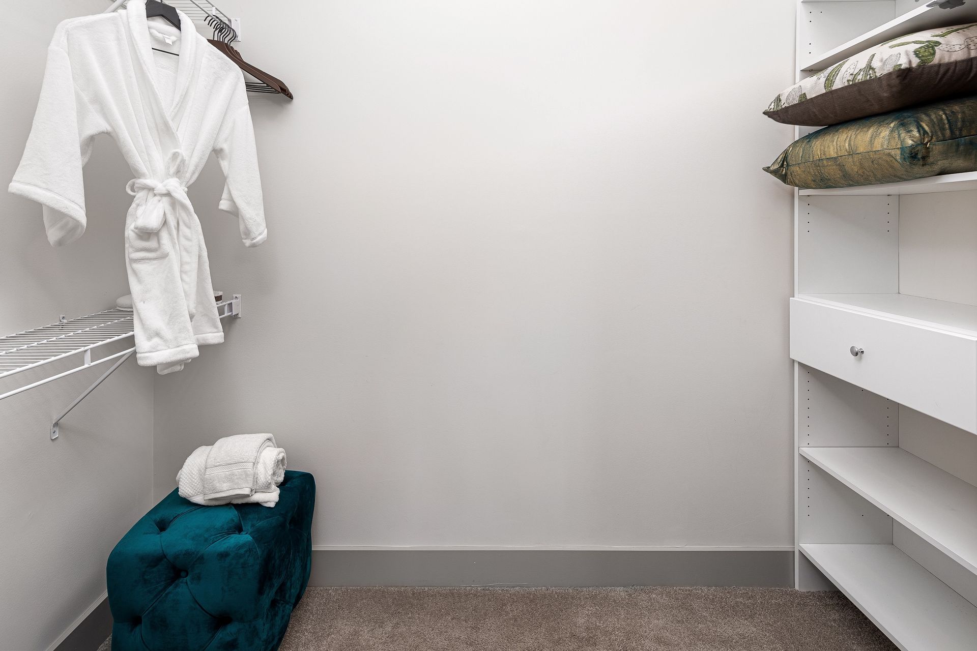 A walk in closet with a robe hanging on the wall and a blue ottoman.