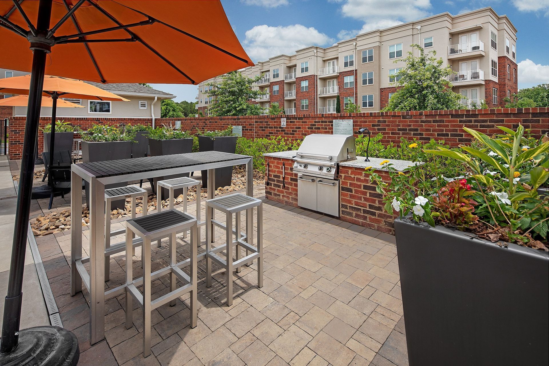A patio with a table and stools under an orange umbrella and a grill.