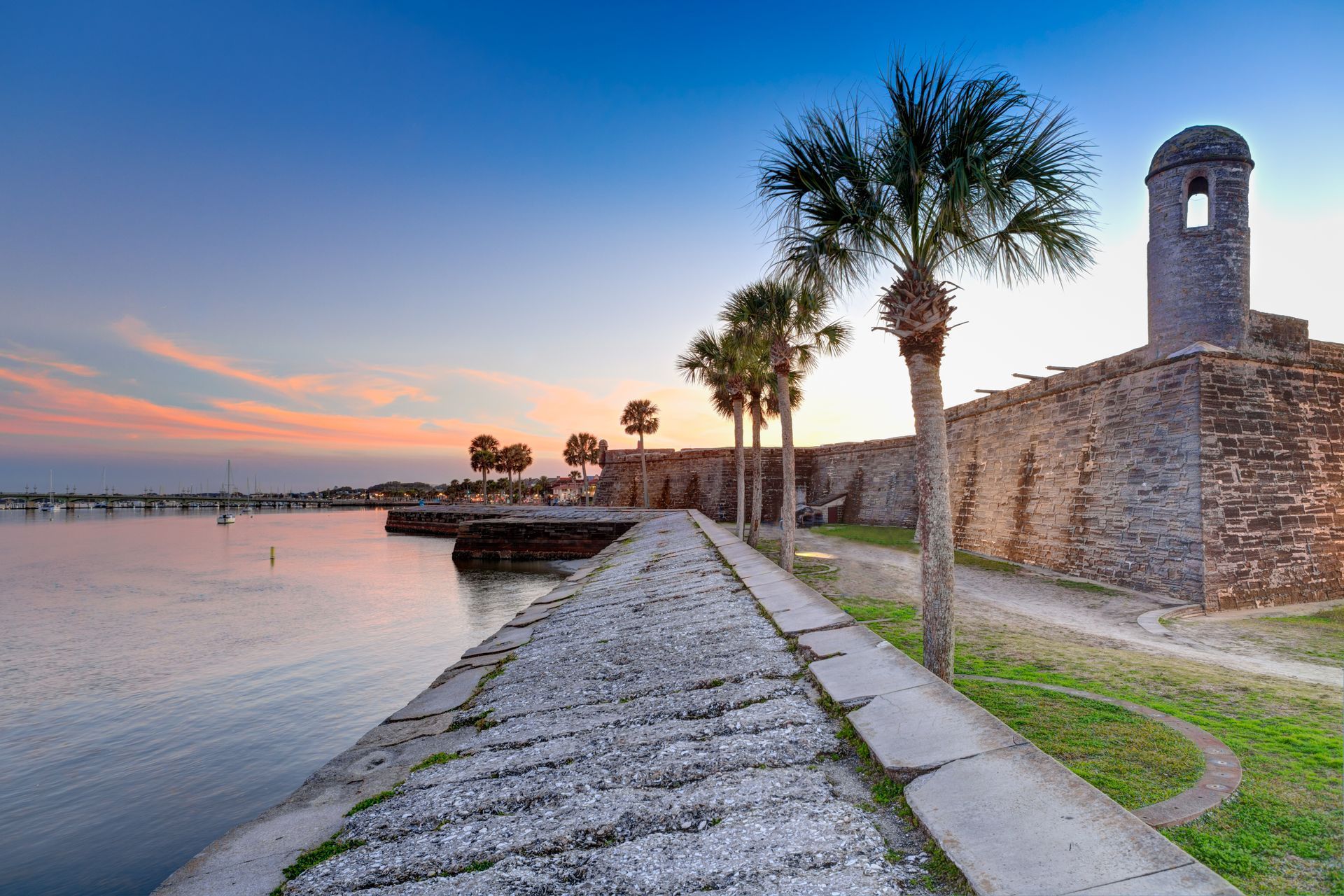 A stone walkway leading to a brick building next to a body of water with palm trees.