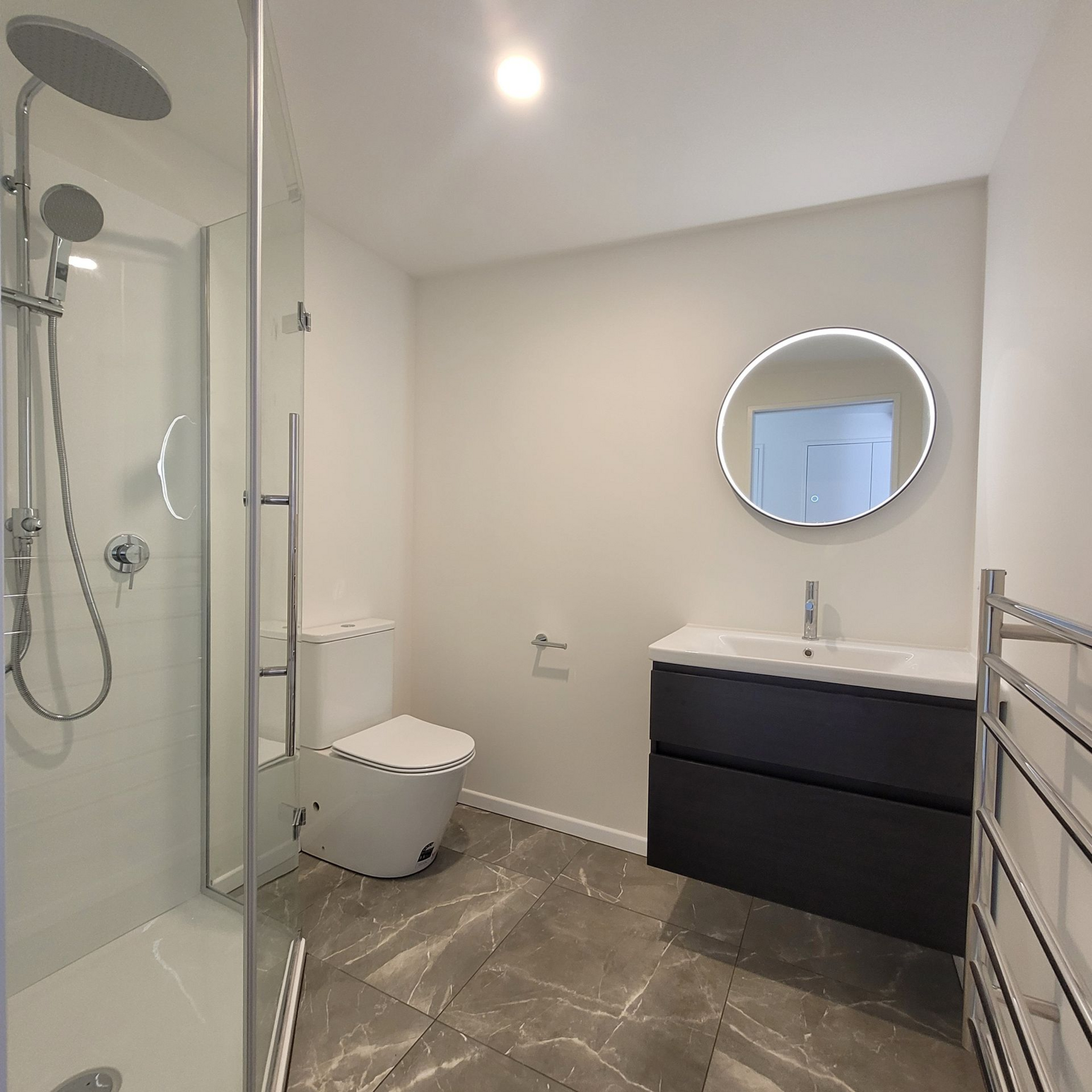 Modern bathroom featuring a glass-enclosed shower, toilet, dark grey vanity with a round LED mirror, and tiled floors.