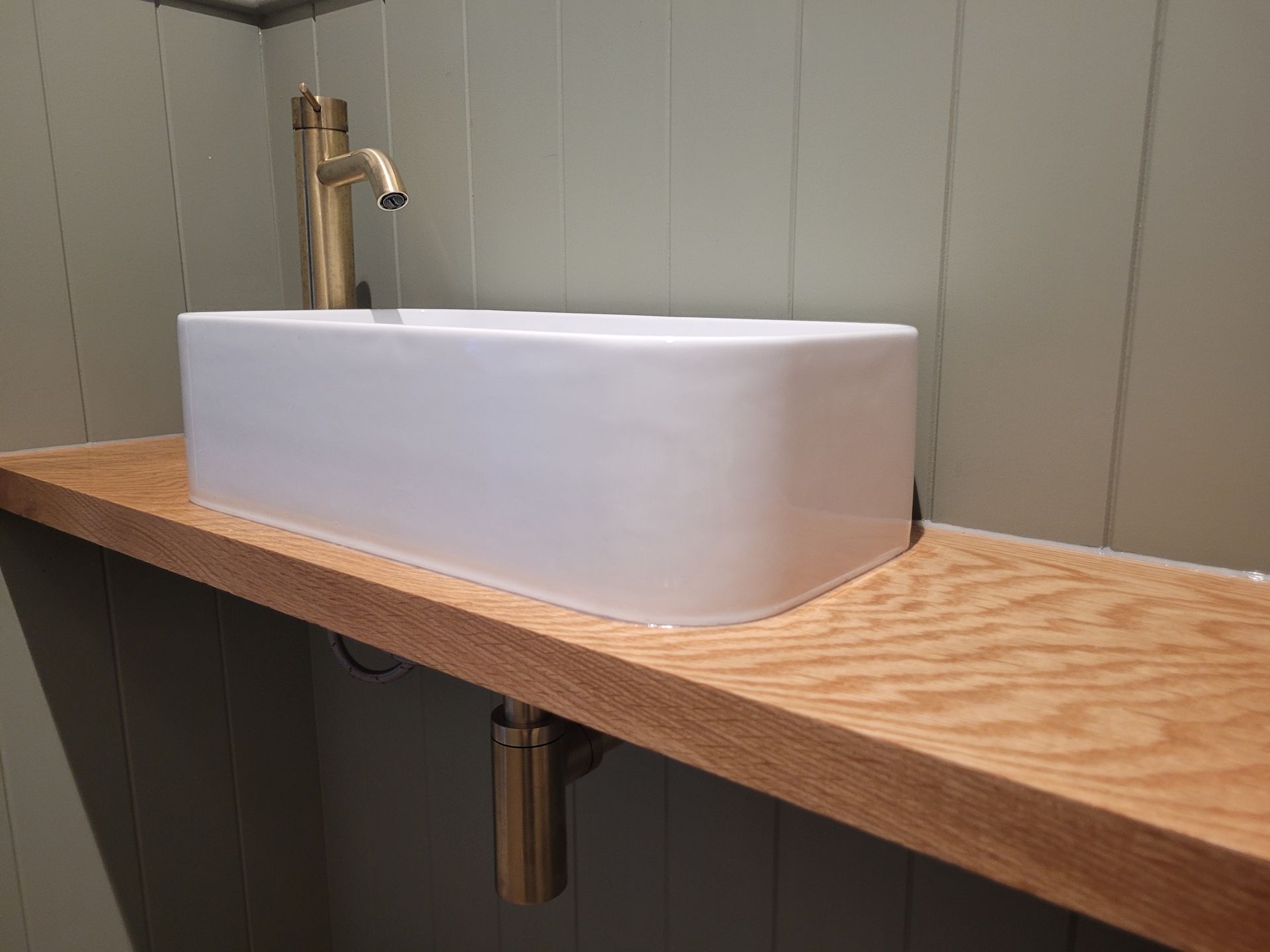 A white, rectangular vessel sink on a natural wood countertop with a brushed gold faucet and matching drain pipe.