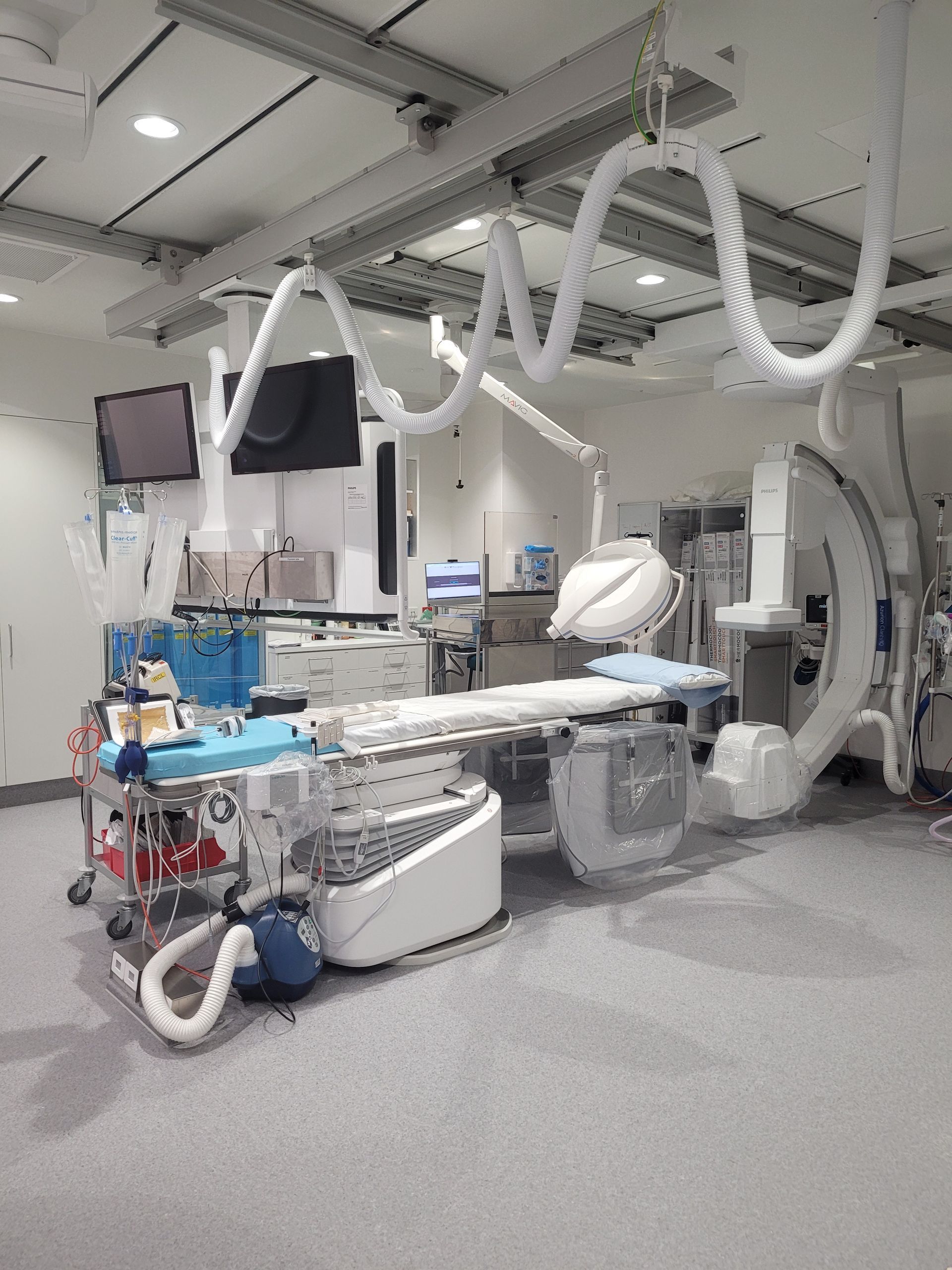 A medical operating room featuring a surgical table, a C-arm X-ray machine, and various clinical monitors and equipment.