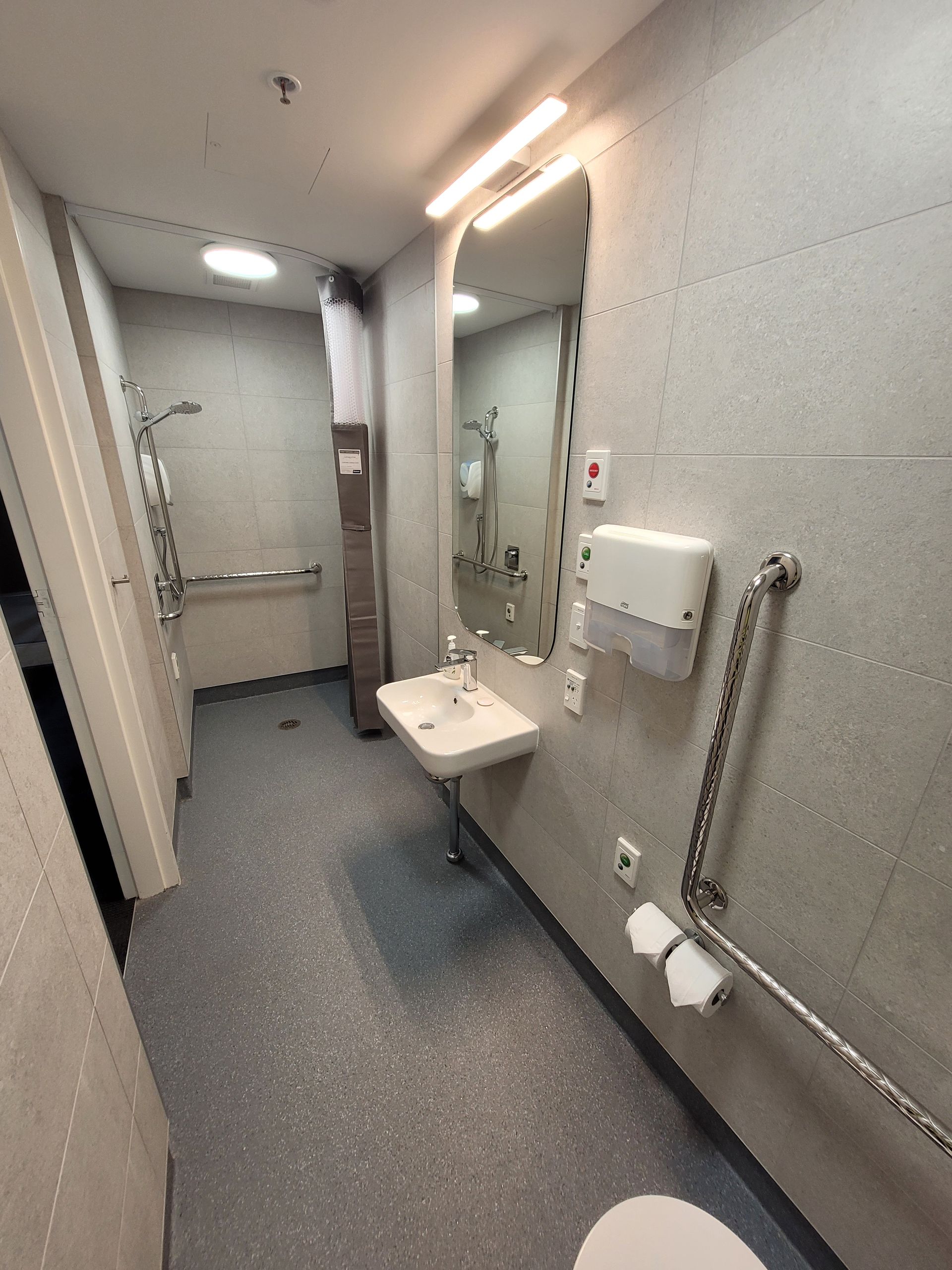A modern, accessible bathroom with a sink, mirror, wall-mounted soap dispenser, and grab bars near the toilet.