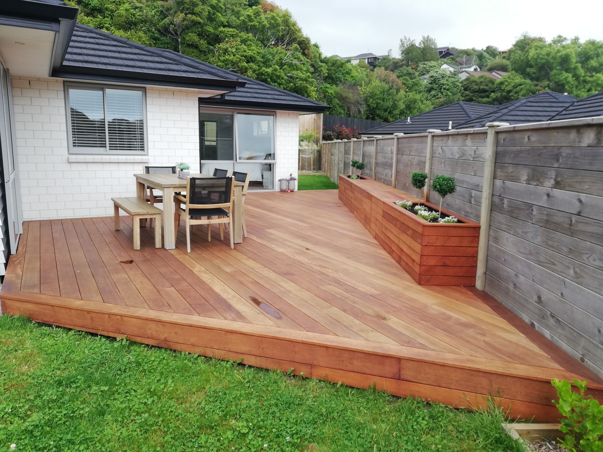 A wooden deck attached to a house with a dining set and a built-in planter box against a tall wooden fence.