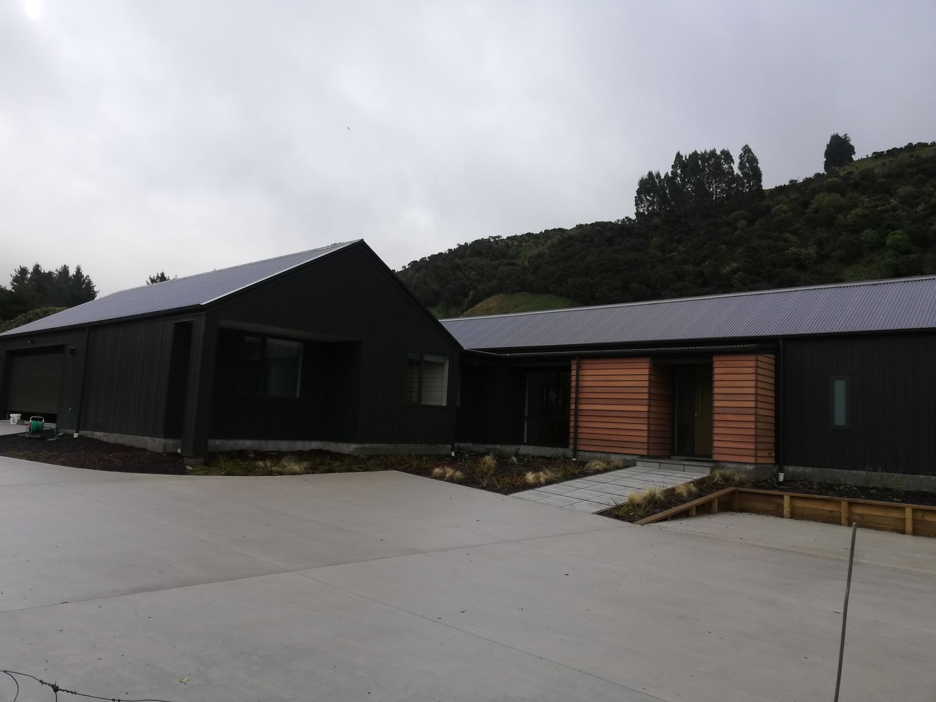 A modern house with dark siding and wood accents, featuring a large concrete driveway set against a forested hillside.