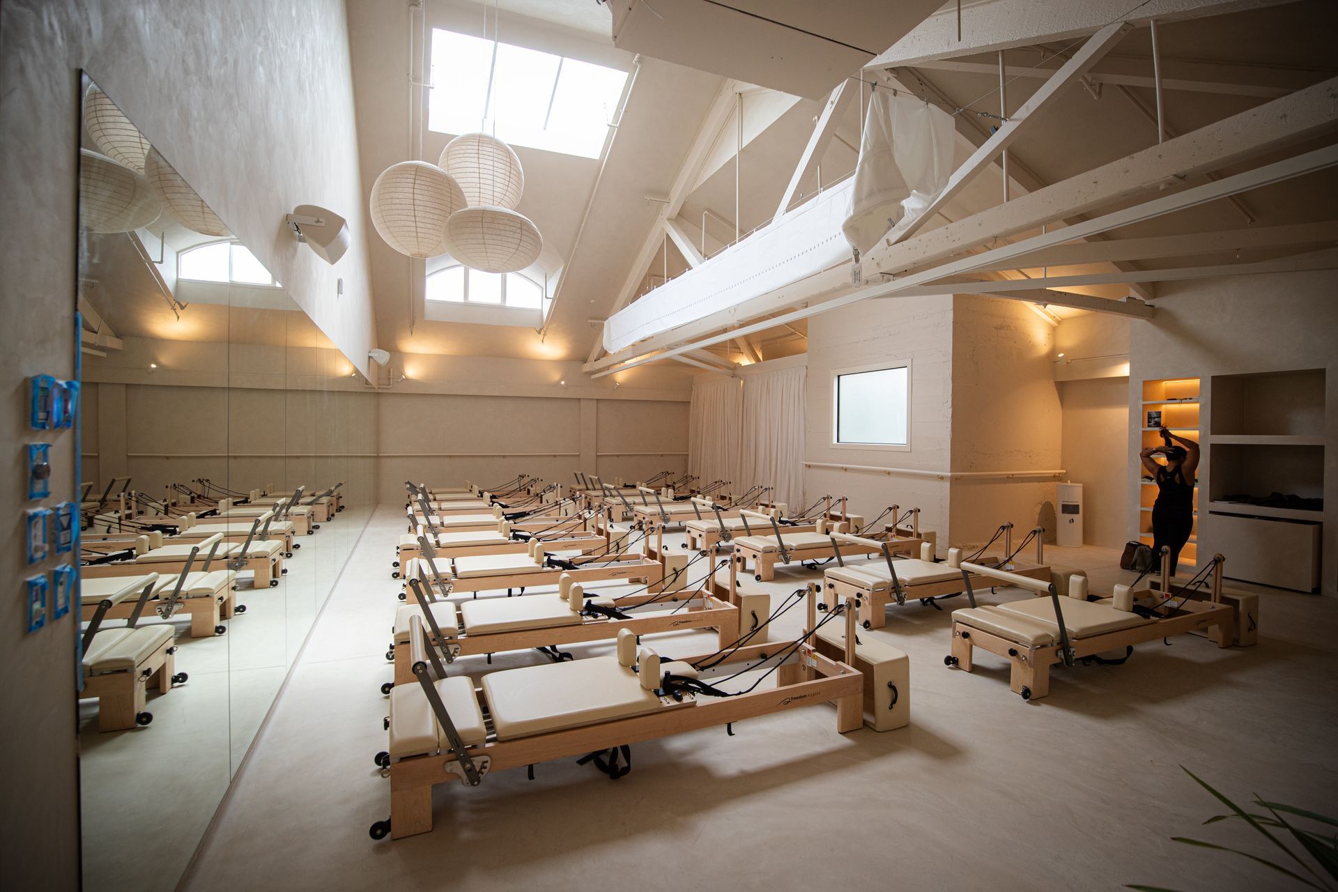 A bright, minimalist studio filled with rows of light-wood Pilates reformers, with a mirror wall and high vaulted ceilings.