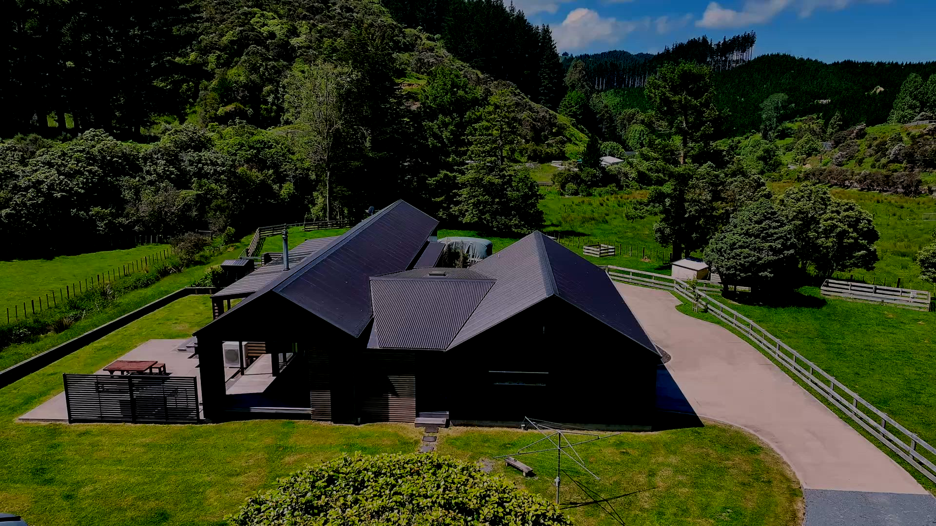 An aerial view of a dark-roofed house set within a lush, green rural landscape with trees and a paved driveway.