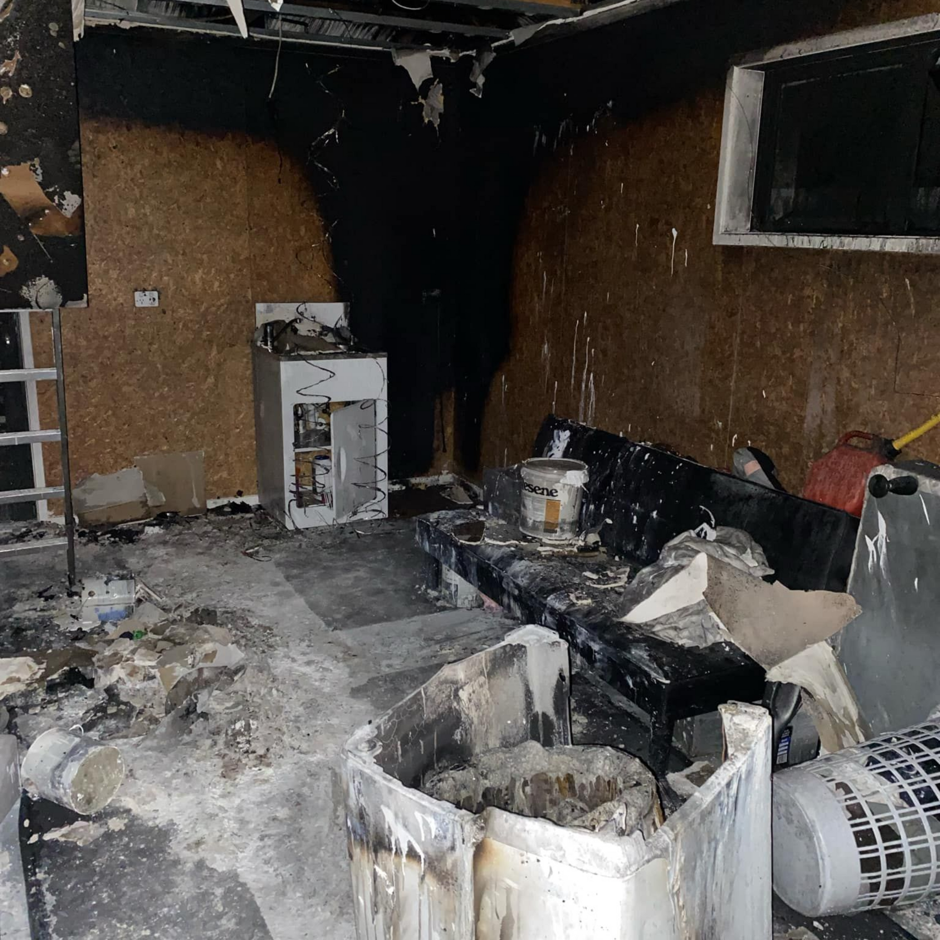 A room damaged by fire, featuring charred walls, debris on the floor, a damaged sofa, and remnants of furniture.