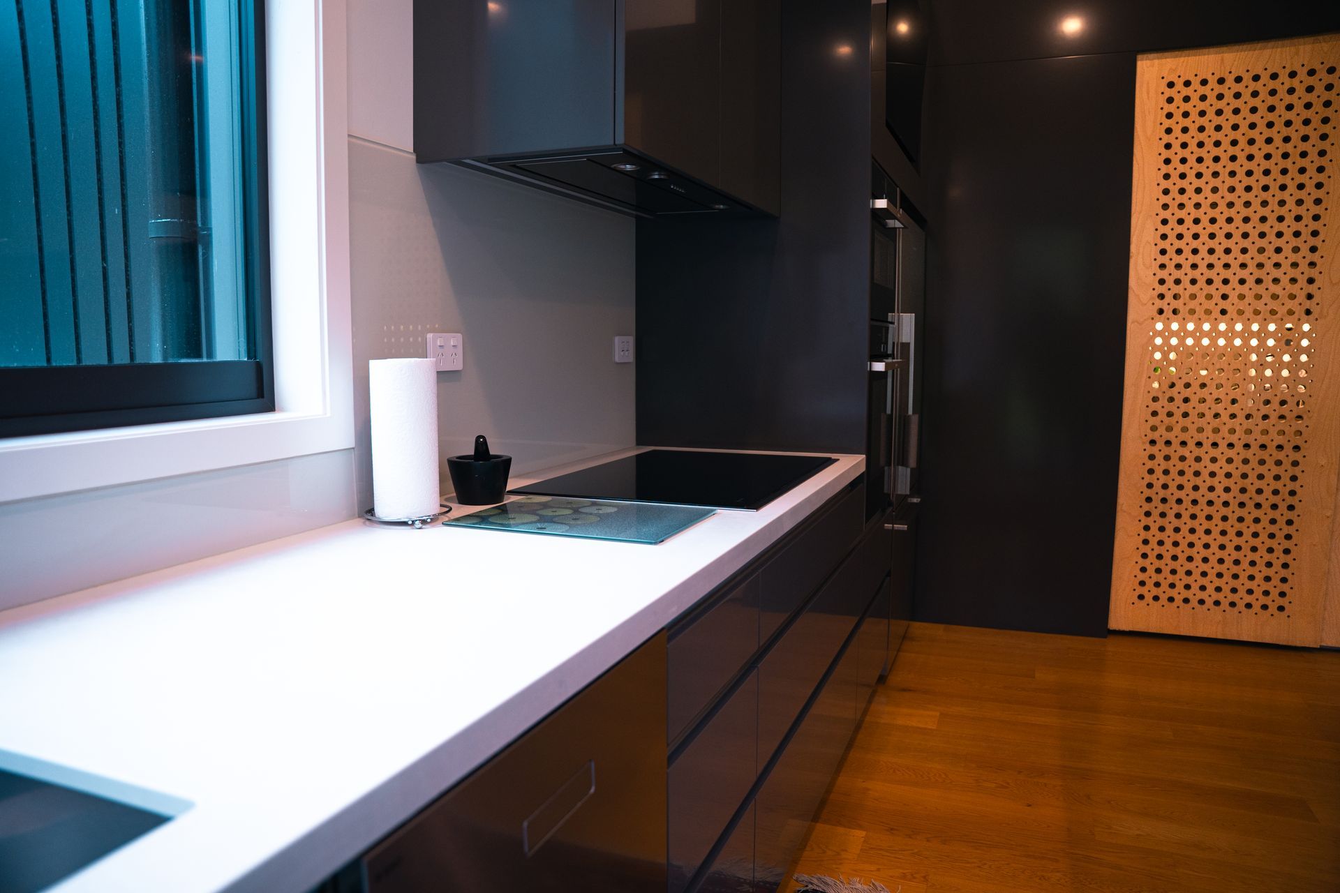 A modern kitchen with dark cabinets, a white countertop, a cooktop, a paper towel roll, and a patterned wooden door.