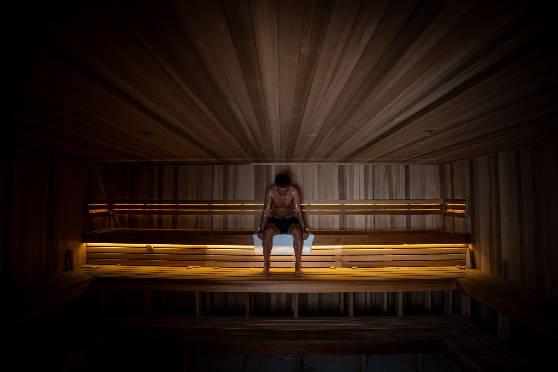 A lone figure sits with bowed head on a bench inside a dimly lit, wood-paneled sauna illuminated by ambient light.