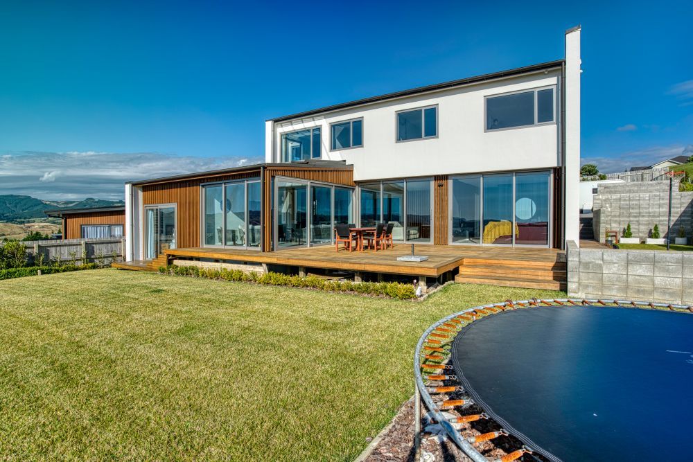 A modern two-story home with a wooden deck and large glass doors, seen from a grassy lawn with a trampoline.