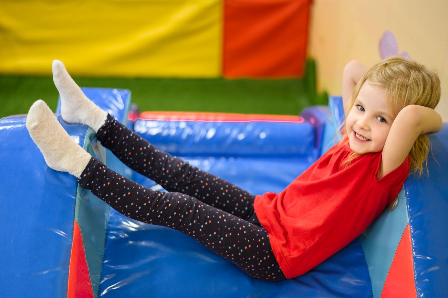 Girl with blonde hair, red shirt, and black leggings smiles, relaxing on blue inflatable in play area.