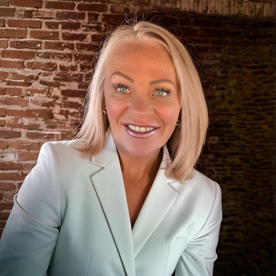 Woman with blonde hair smiles in light blue suit, brick wall backdrop.