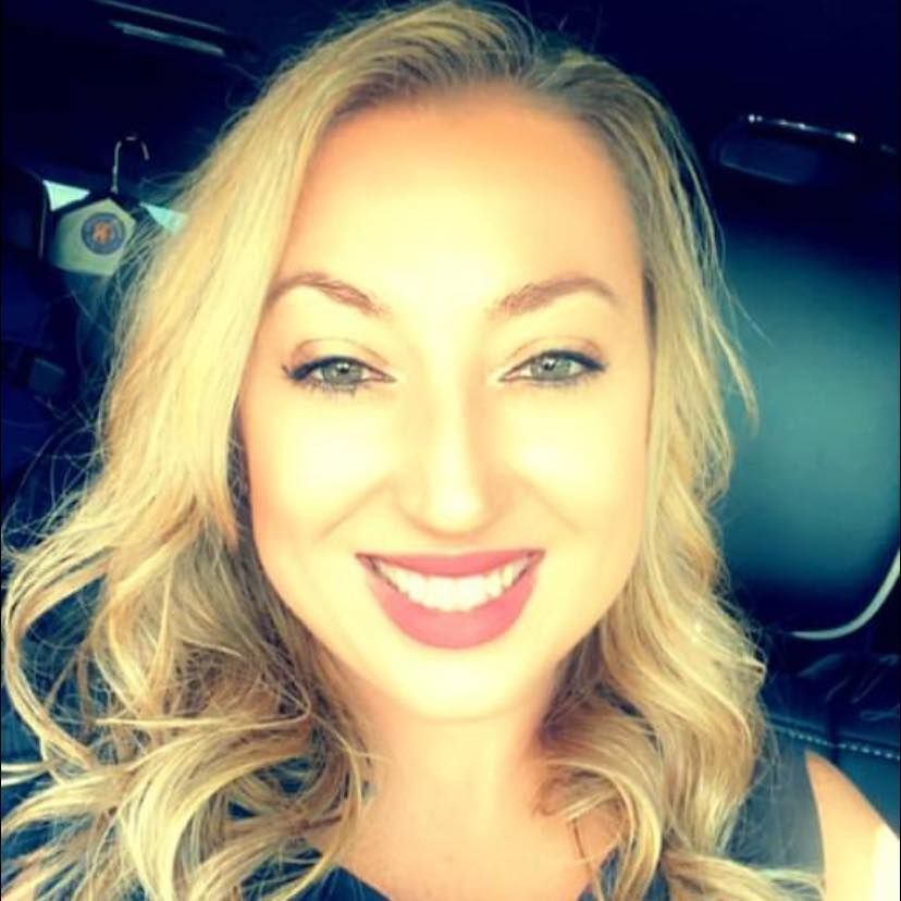 Blonde woman with a big smile, light eyes, and red lipstick; car interior visible.