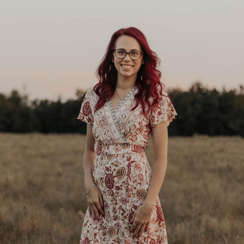Woman with red hair, glasses, in a floral dress, smiling in a field.