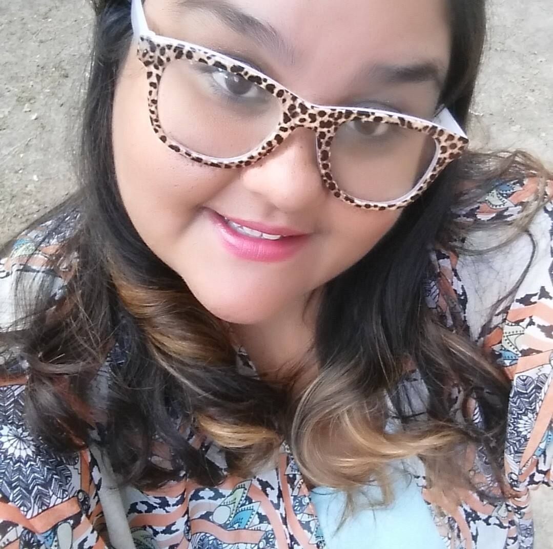 Woman with glasses smiling outdoors. Leopard print frames, brown hair, patterned top.