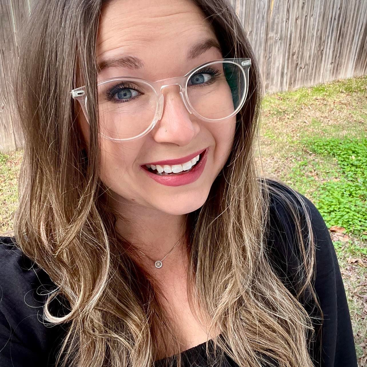 Woman with glasses smiling outside. Blonde hair, black shirt, necklace.