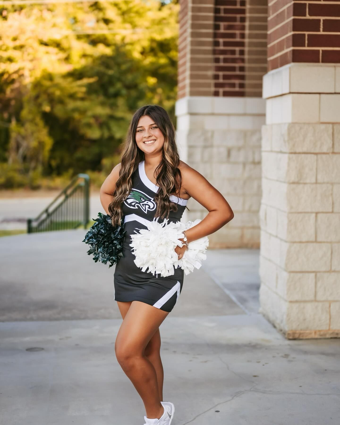 Cheerleader smiling, holding pom-poms, wearing gray uniform, standing outside near brick building.