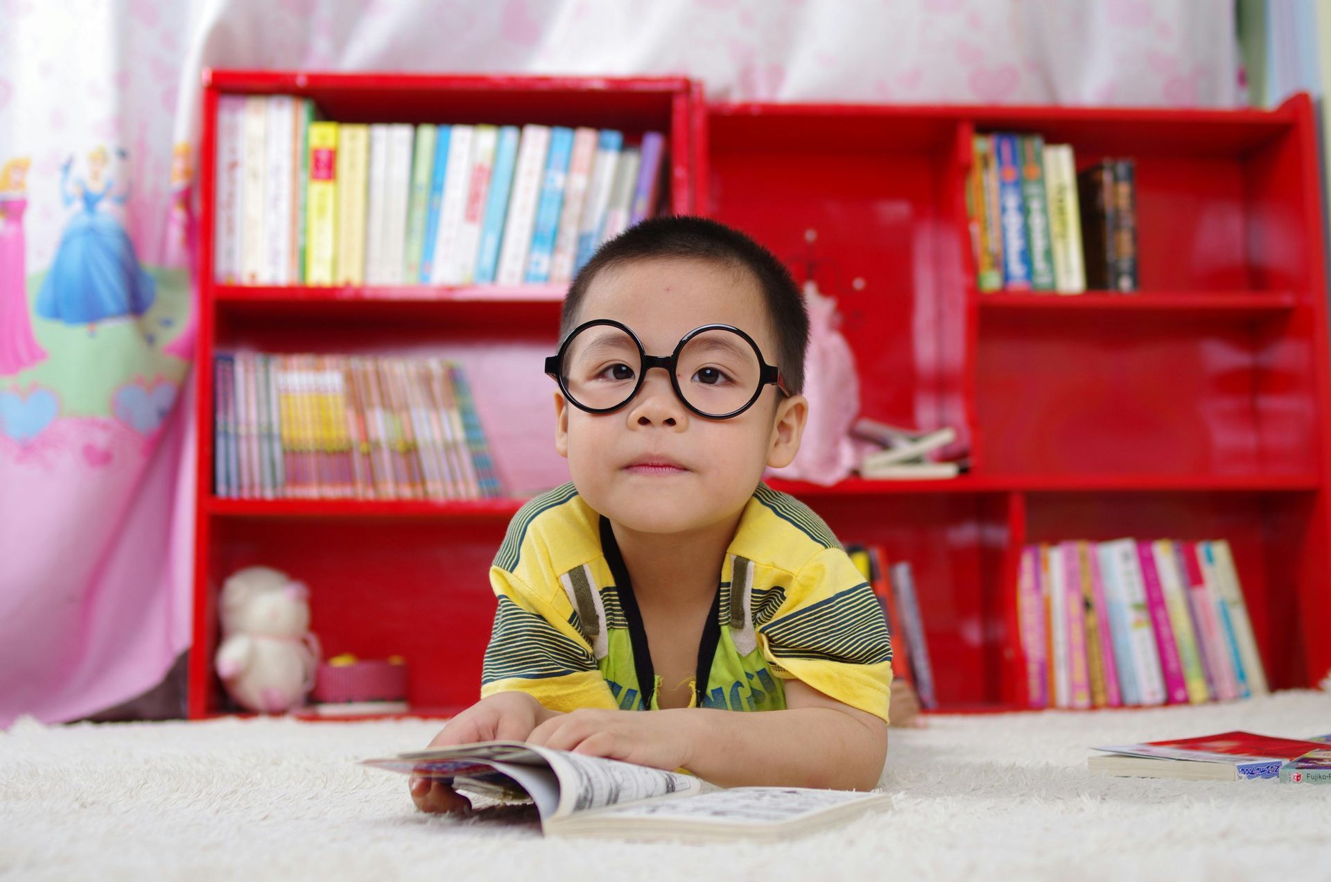Boy with glasses reading in front of a red bookshelf.