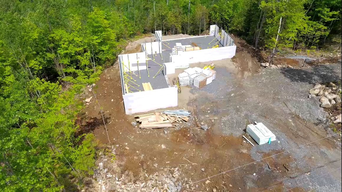 Chantier de construction avec des murs de fondation blancs entourés de terre, d'arbres et de matériaux de construction.