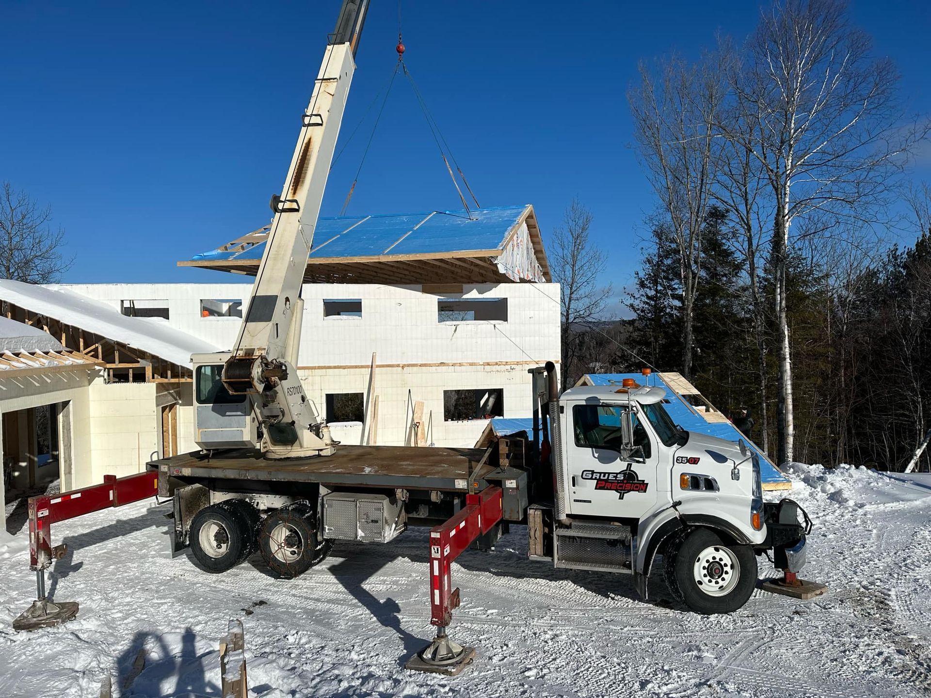 Camion-grue soulevant un panneau mural pour le poser sur une maison en construction dans un décor enneigé.