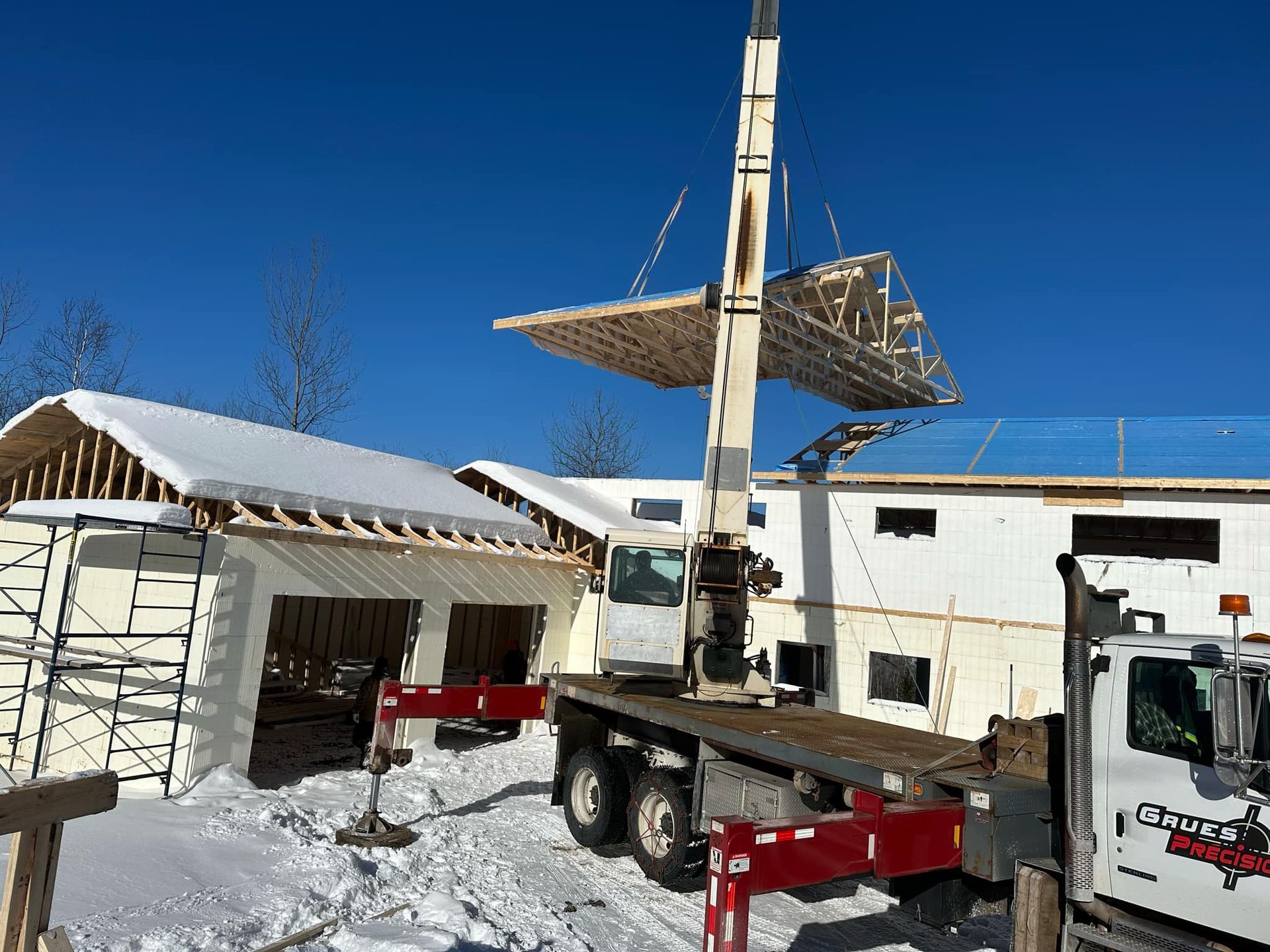 Grue soulevant une charpente de toit pour la poser sur un bâtiment en construction, décor enneigé, ciel d'un bleu éclatant.