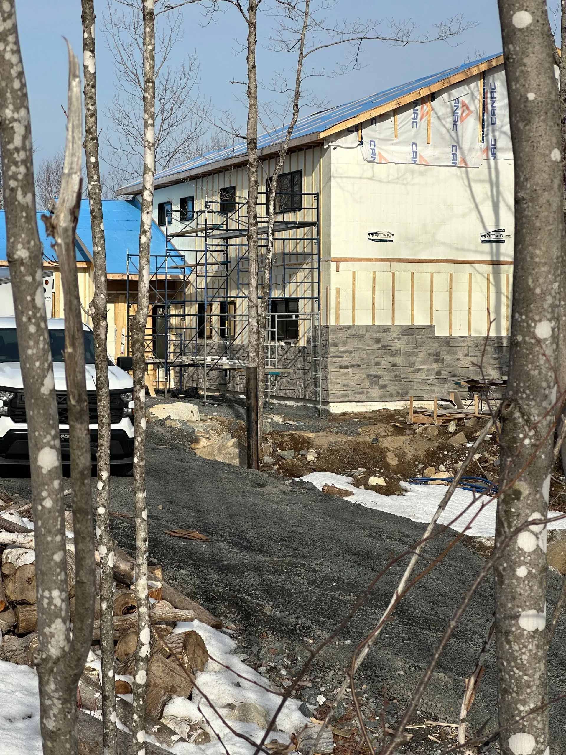 Chantier de construction d'un bâtiment avec ossature bois apparente, isolation et bardage extérieur partiellement installé. Allée de gravier et arbres au premier plan.