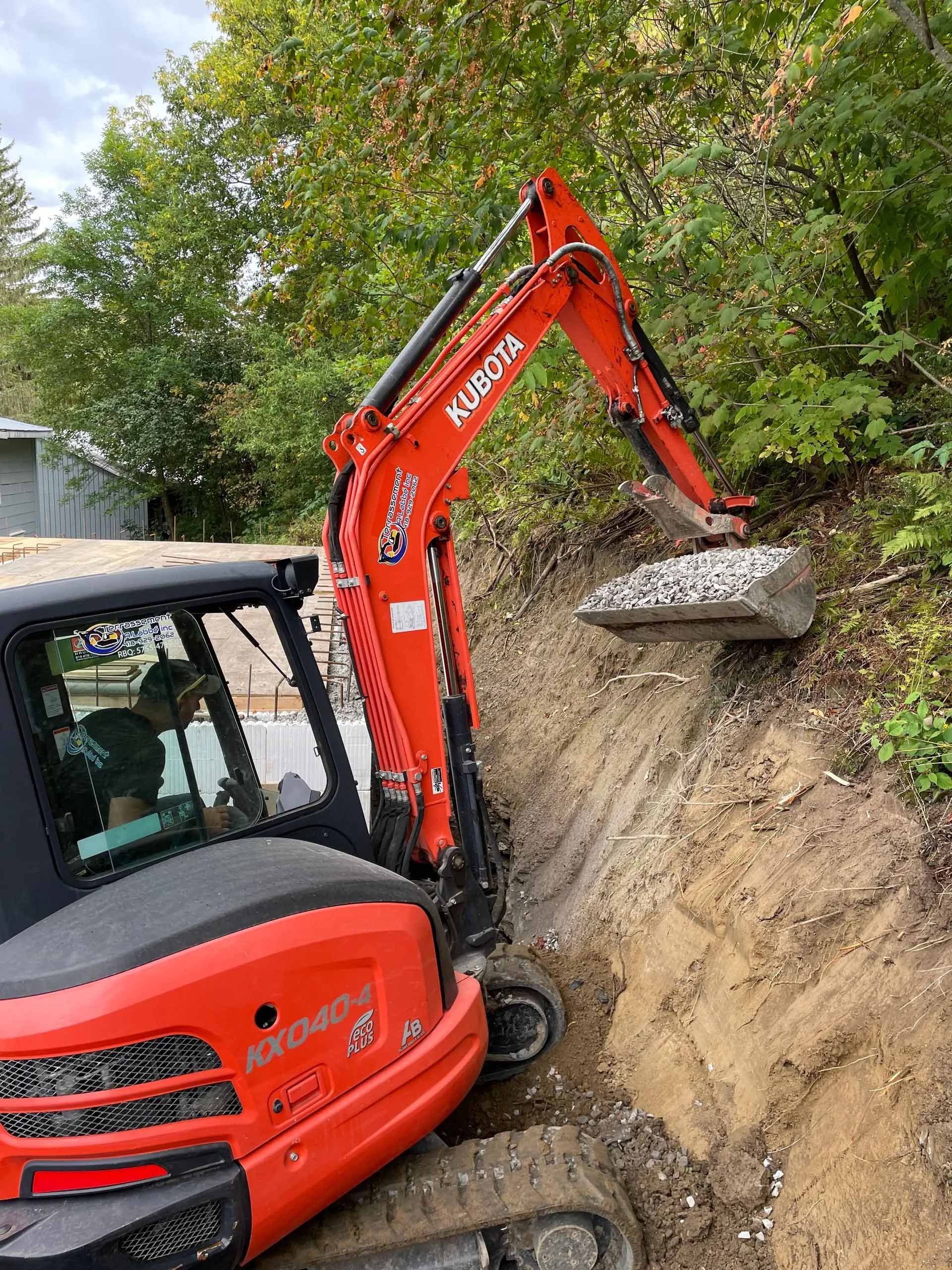 Une pelle mécanique Kubota orange creuse et soulève un gros rocher d'un fossé.