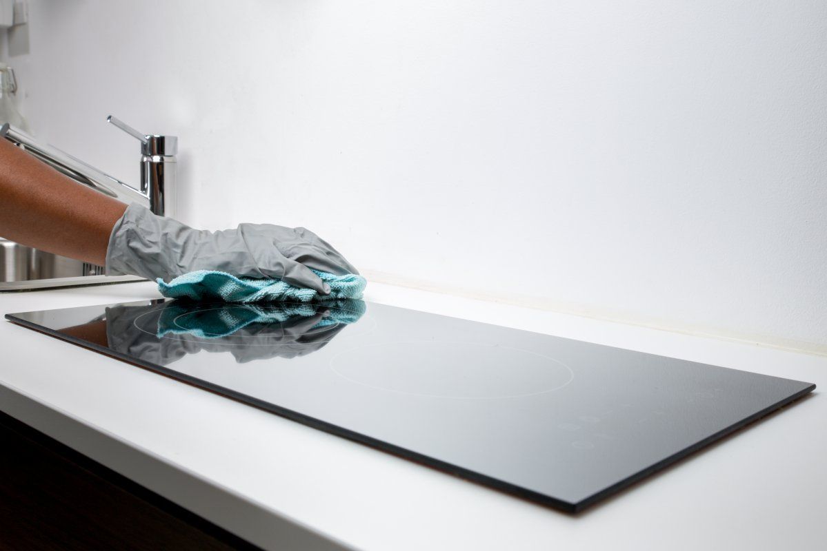 Person wearing a glove wiping a black stovetop with a blue cloth in a white kitchen.