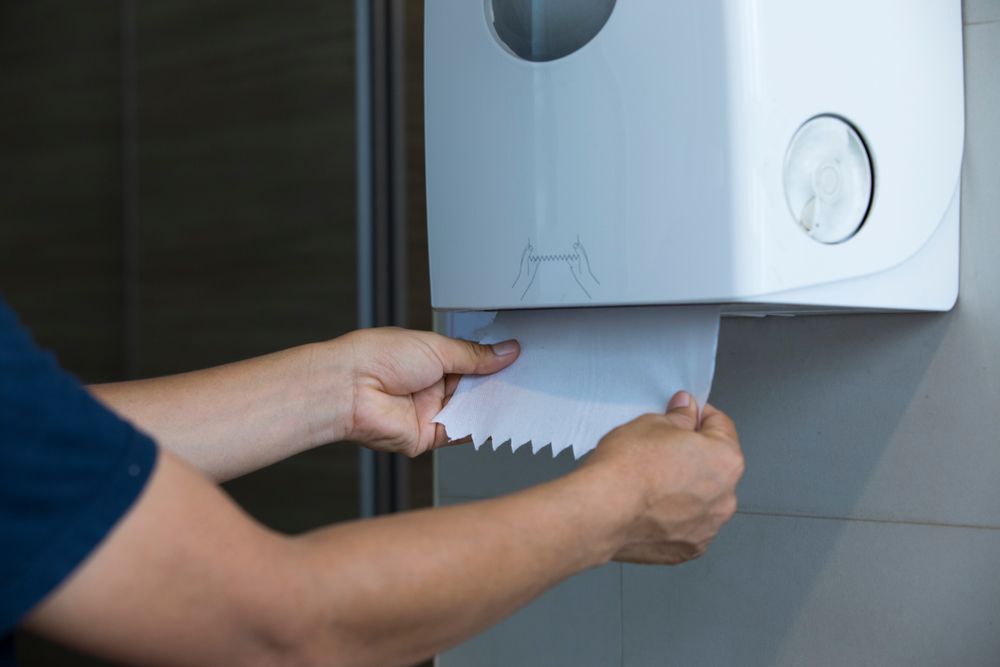 Person pulling a paper towel from a white dispenser mounted on a wall.