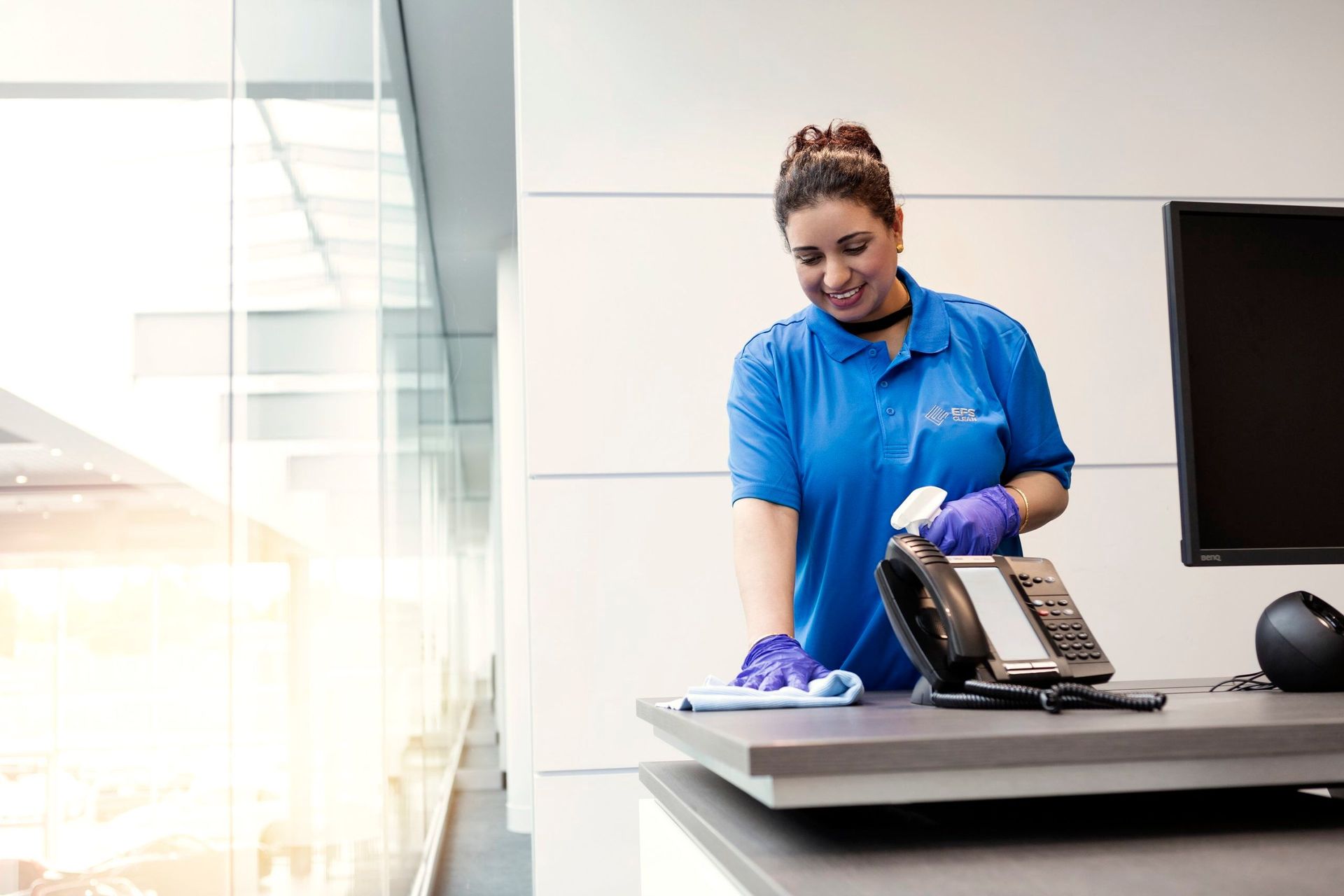 Woman in blue uniform cleaning a desk with spray and cloth. Office setting, large window.