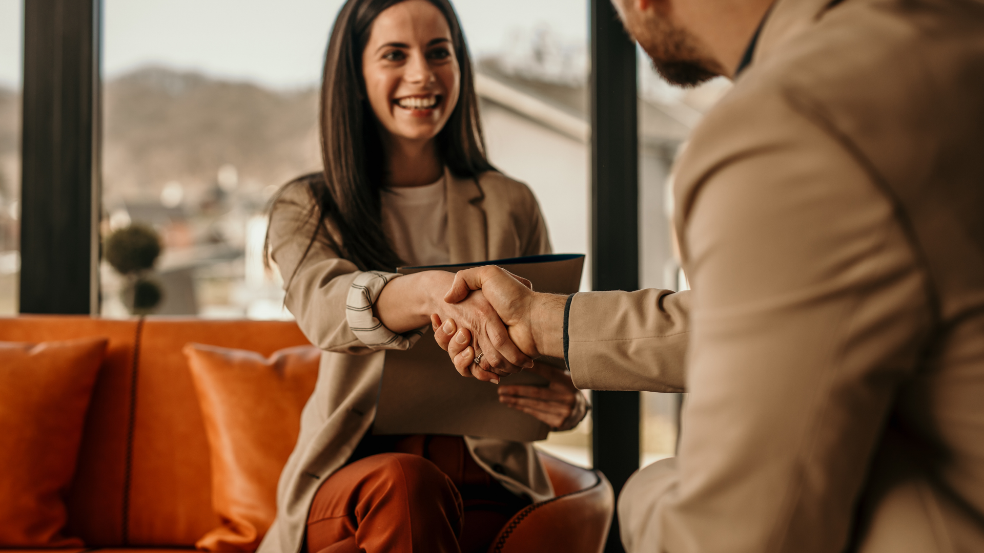 Two people shaking hands across a table in a bright lounge with orange seating.
