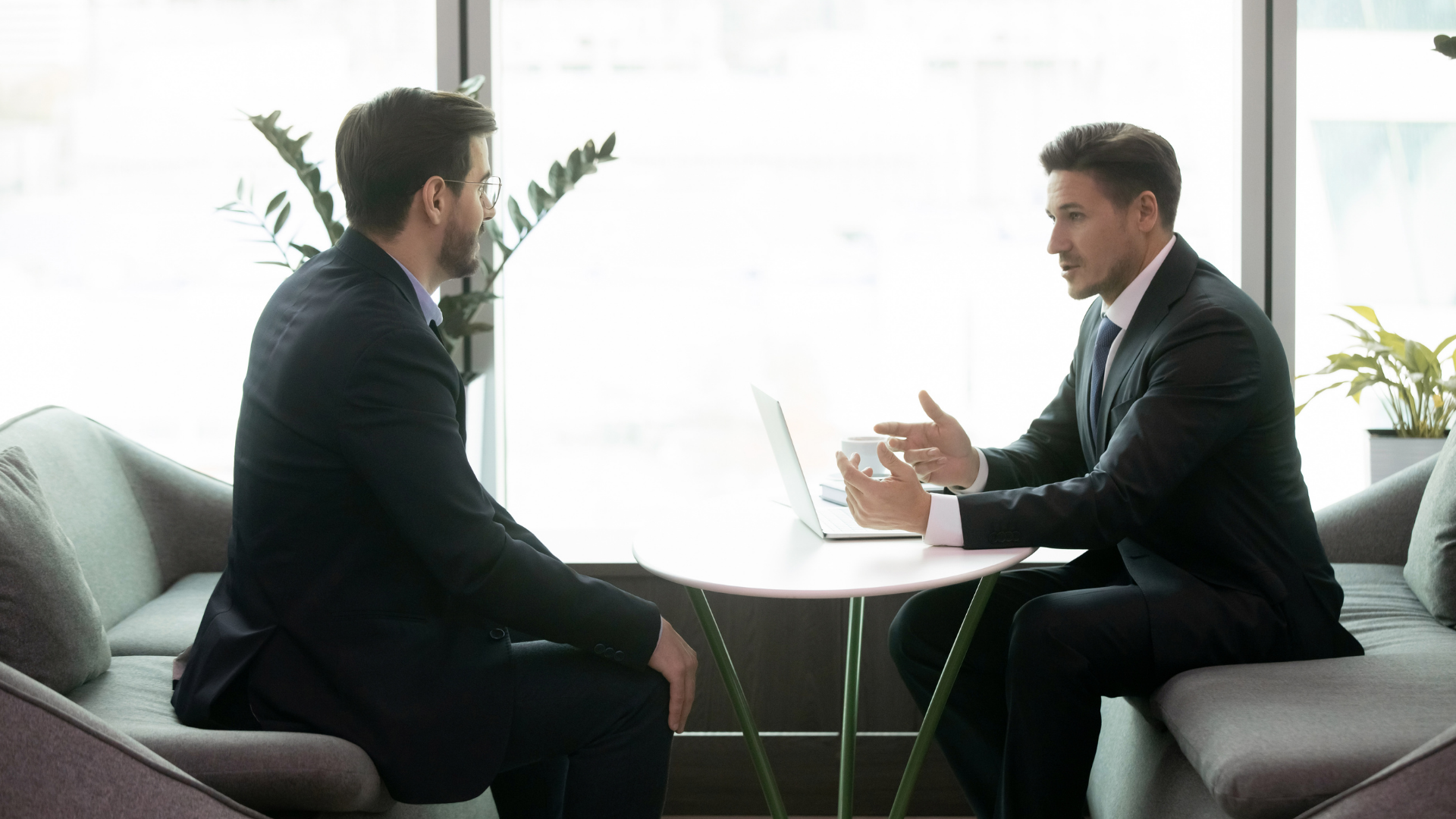 Two professionals in suits discuss business across a small table in a bright office lounge.