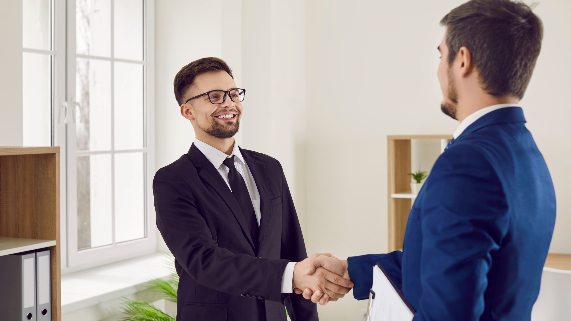 Two men in suits are shaking hands in an office.