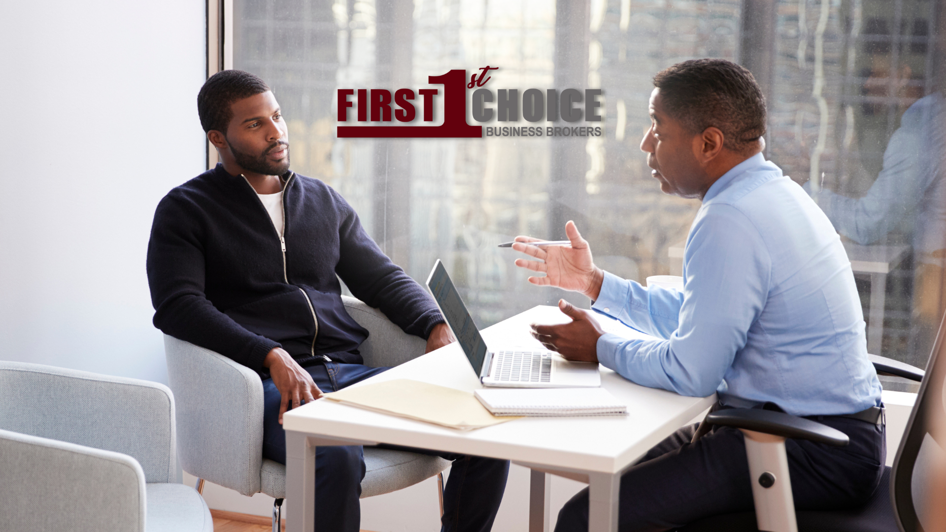 Two men are sitting at a table with a laptop and talking to each other.