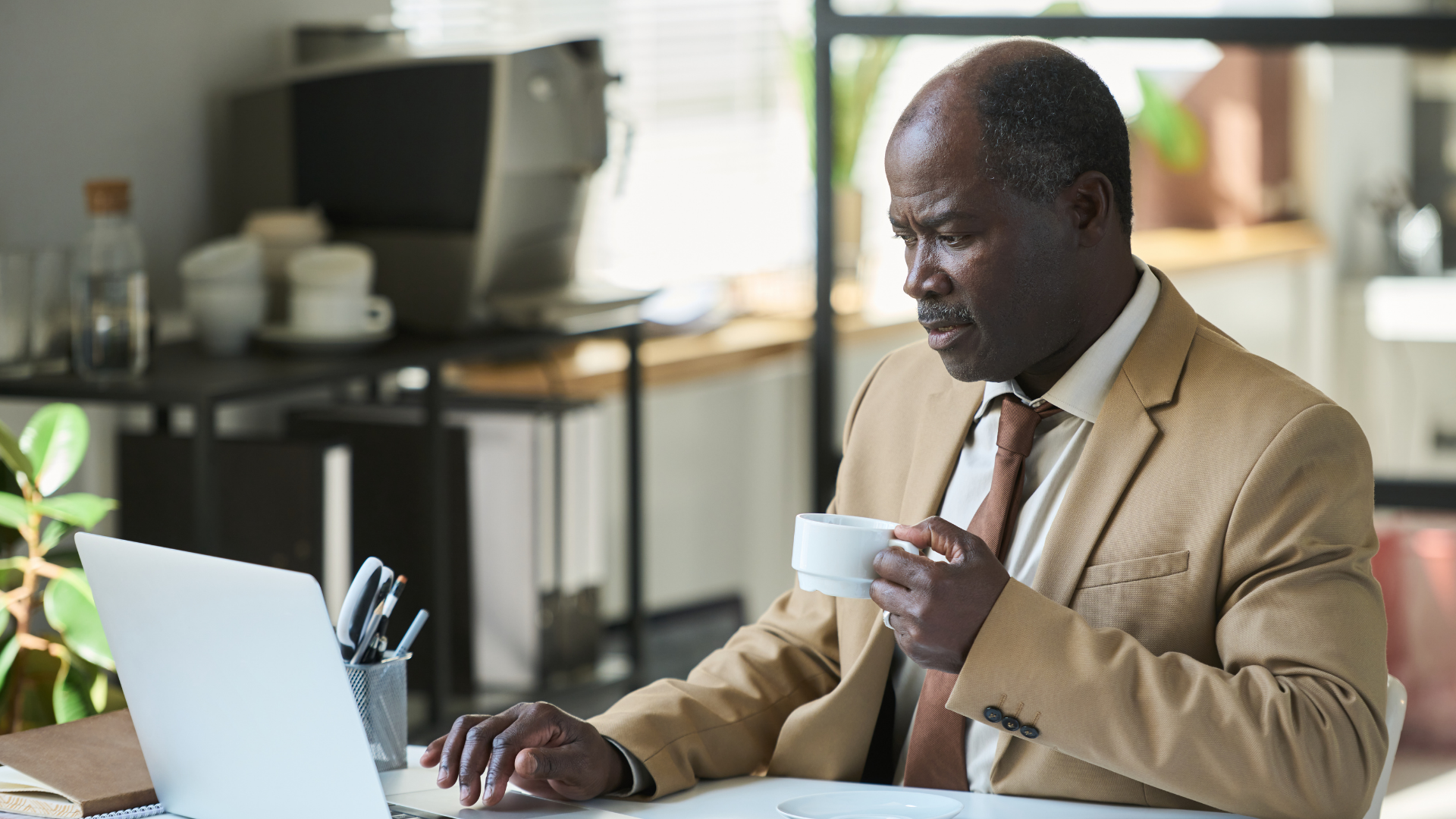 Man in tan suit, holding a cup, looking at laptop in an office setting.