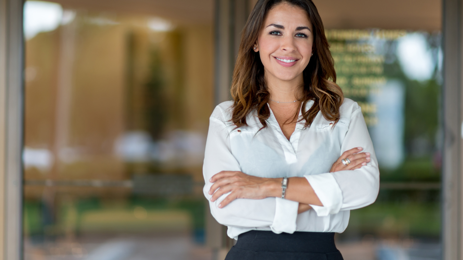 A woman in a white shirt and black skirt is standing with her arms crossed and smiling.