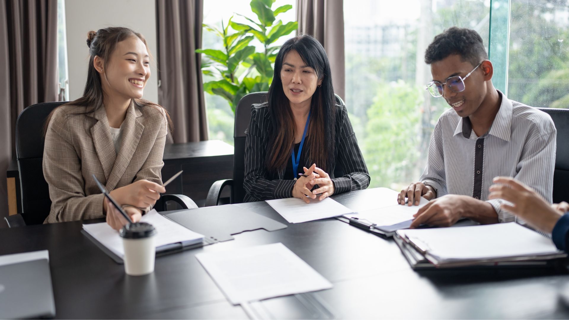 Three coworkers in a meeting at a conference table, discussing papers and a laptop.