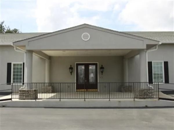 Gray building with porch and black iron railing. Double doors centered, flanked by windows and sconces.
