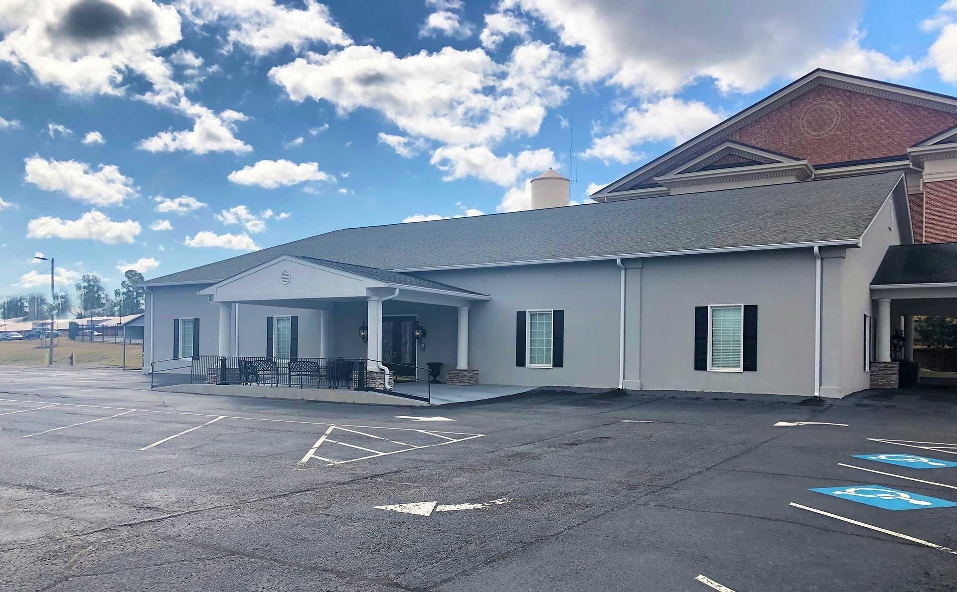 Gray building with a covered entrance, black shutters, and a parking lot under a cloudy sky.