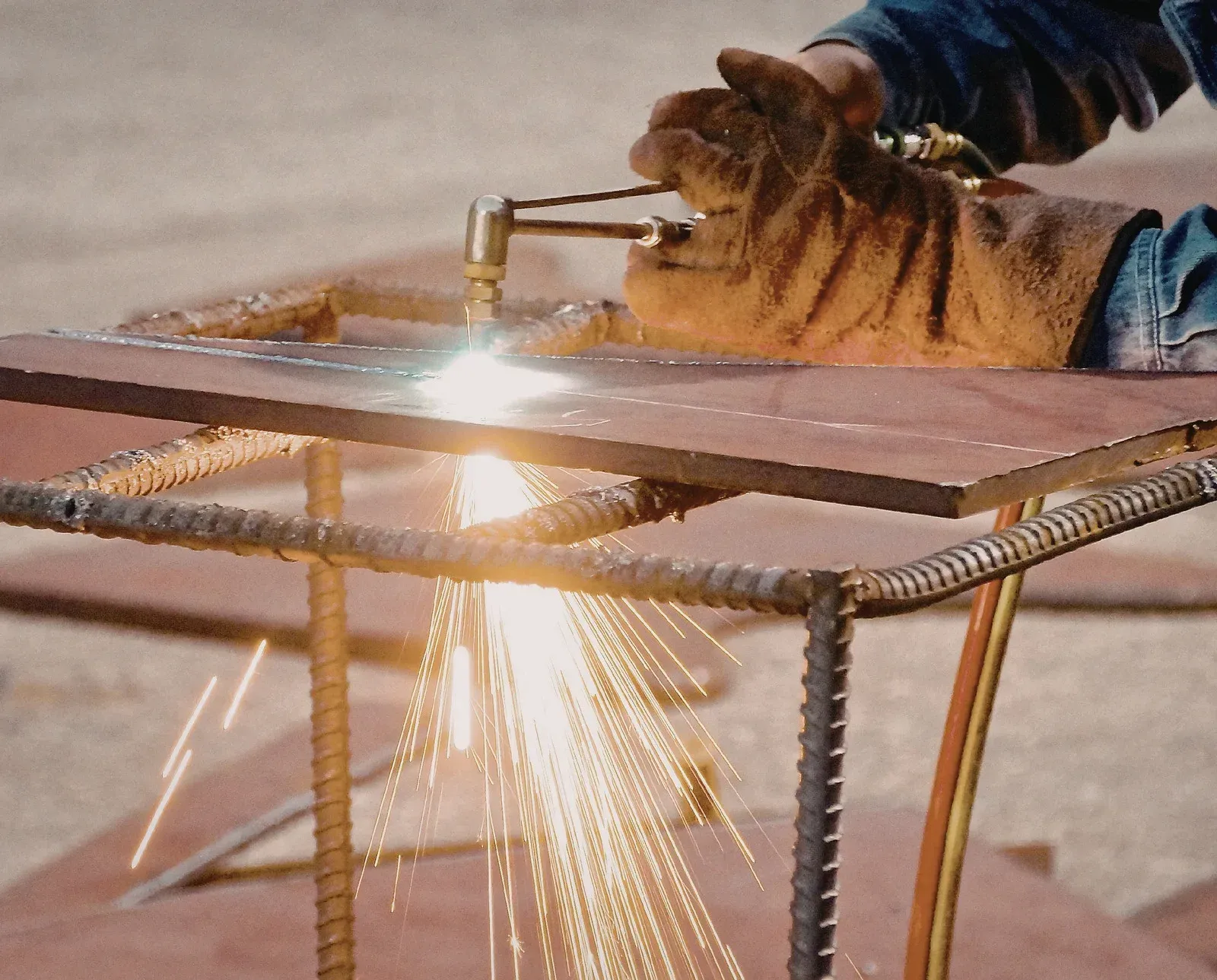 A person wearing leather work gloves uses a torch to cut through a thick metal plate supported by a rebar frame.