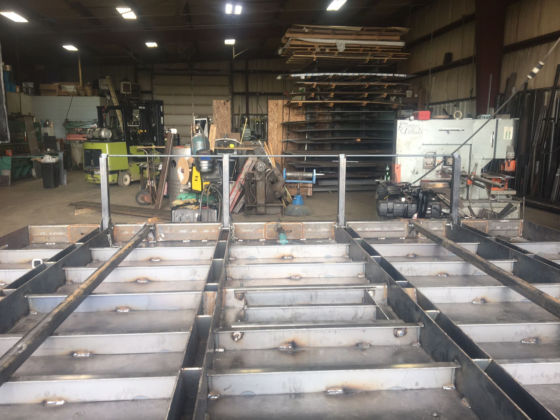 A workshop floor view shows a large, rectangular metal frame structure under construction, with industrial tools behind.