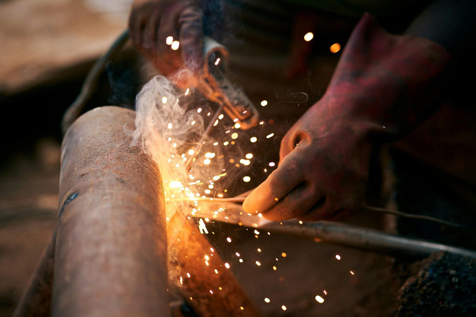 Close-up of a worker in protective gloves performing metal welding, creating bright sparks and smoke on a metal pipe.