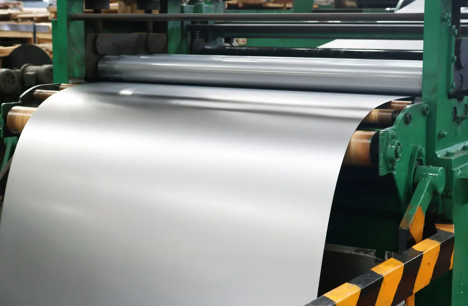 A large, smooth sheet of silver metal feeds through industrial green rollers in a factory setting.