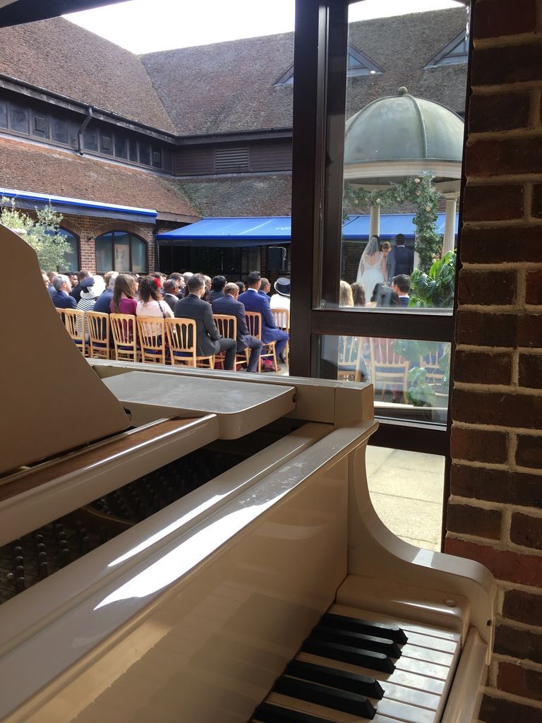 A white grand piano inside with doors open to the outside overlooking a wedding ceremony