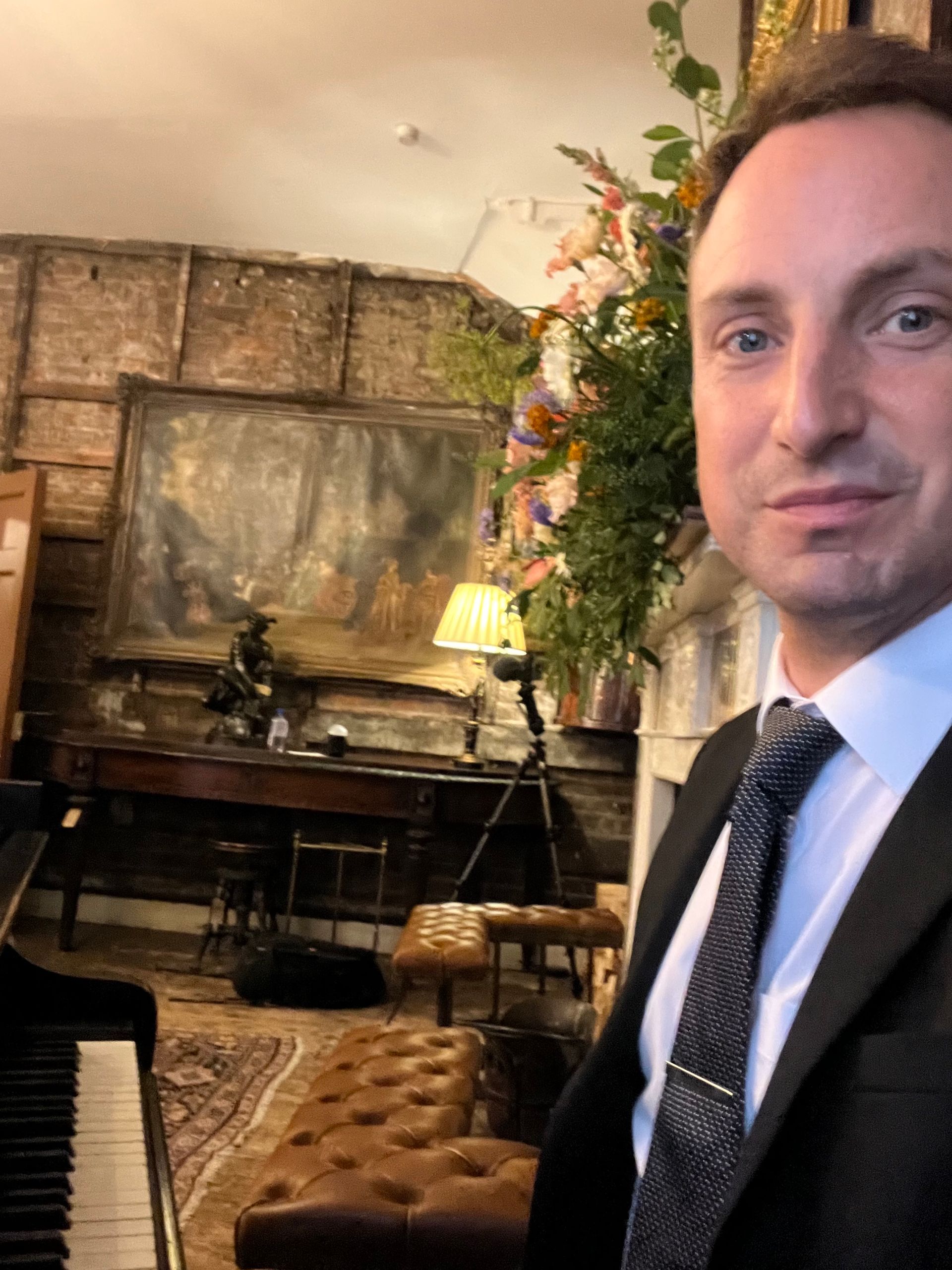 A selfie of a man in a suit sitting by a piano in an old stately home