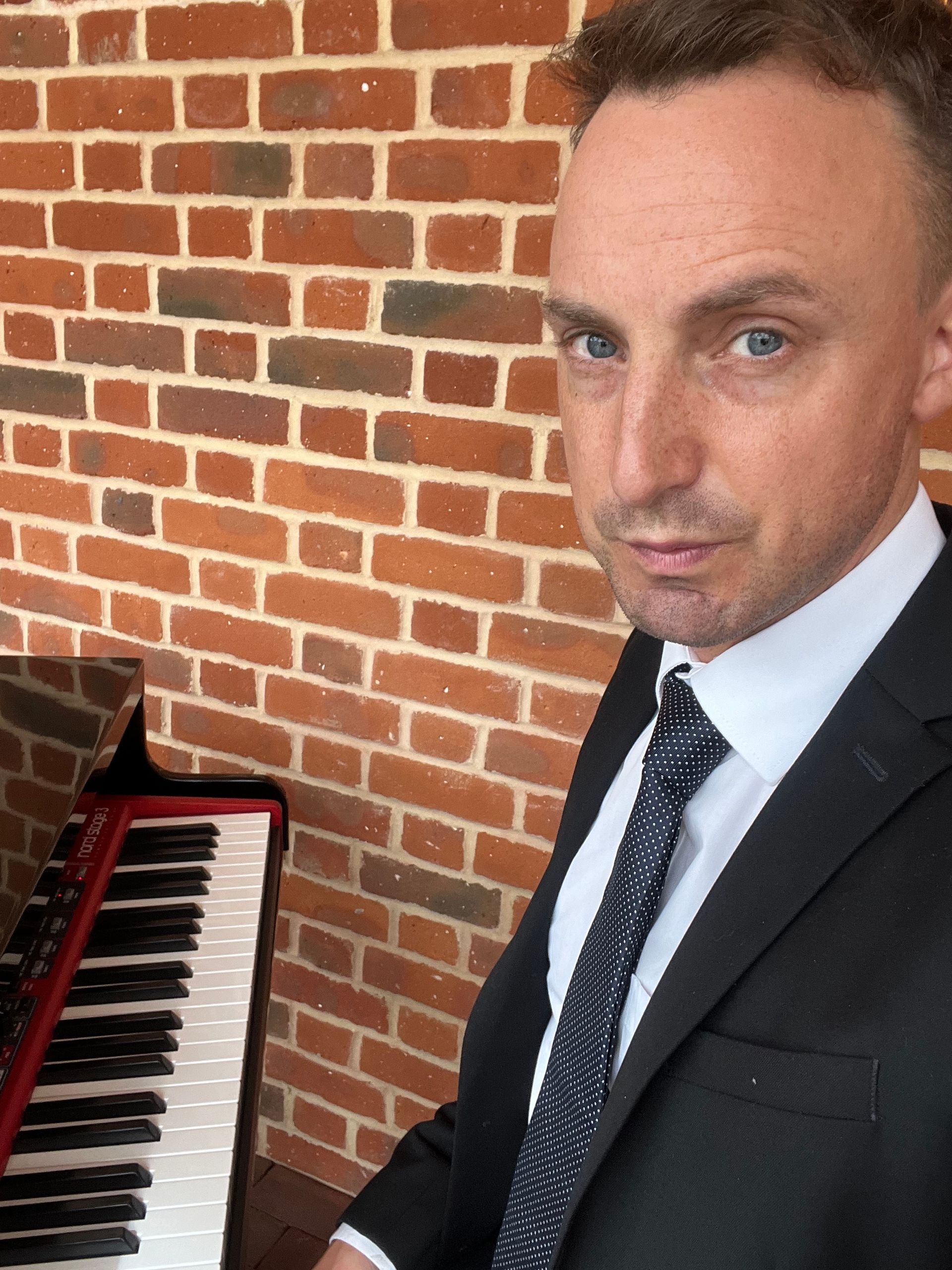 A man in a suits sits by a piano against an exposed brick wall